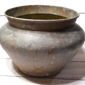 May include: A round, wide-mouthed metal pot with a weathered, tarnished surface showing shades of brown, gray, and hints of green oxidation. The vessel features a flared rim and a curved, bulbous body that tapers toward a narrow, flat base.