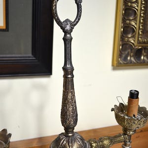 Puede incluir: Una l&aacute;mpara candelabro de metal con un poste central vertical y un asa ovalada en la parte superior, decorada con patrones de hojas. La l&aacute;mpara tiene dos brazos laterales con copas en forma de p&eacute;talos, una con un casquillo de bombilla.