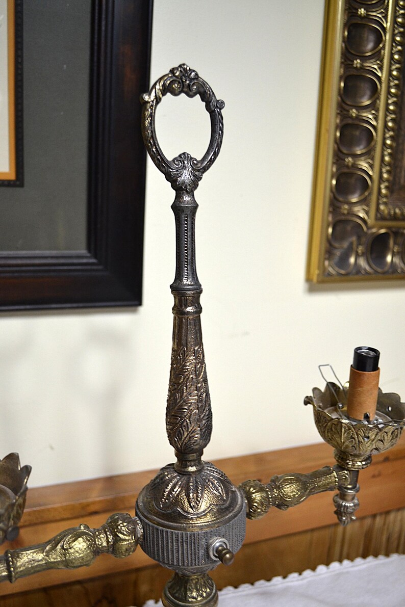 Puede incluir: Una l&aacute;mpara candelabro de metal con un poste central vertical y un asa ovalada en la parte superior, decorada con patrones de hojas. La l&aacute;mpara tiene dos brazos laterales con copas en forma de p&eacute;talos, una con un casquillo de bombilla.