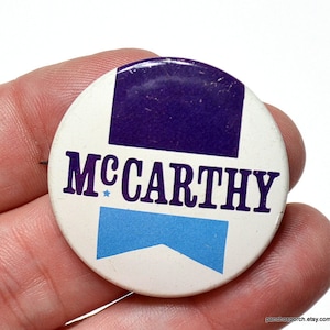 May include: A vintage political campaign button with the name "McCarthy" in bold, dark blue letters. The button has a white background with a purple square at the top and a light blue banner at the bottom. A small blue star is next to the name.