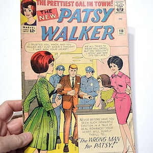 May include: A vintage comic book cover of "The New Patsy Walker" #118. The cover features a colourful illustration of a dramatic scene with several characters, including Patsy Walker, police officers, and a man in a suit. The comic book is held by a hand.
