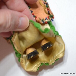 Puede incluir: Primer plano de una falda y zapatos de mu&ntilde;eca vintage. La falda es de color melocot&oacute;n con ribetes verdes y detalles dorados. Los zapatos de la mu&ntilde;eca son de color canela con suelas negras. La falda de la mu&ntilde;eca est&aacute; sujeta por una mano.