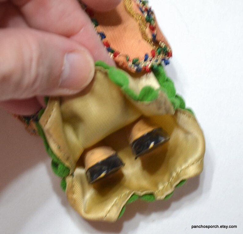 Puede incluir: Primer plano de una falda y zapatos de mu&ntilde;eca vintage. La falda es de color melocot&oacute;n con ribetes verdes y detalles dorados. Los zapatos de la mu&ntilde;eca son de color canela con suelas negras. La falda de la mu&ntilde;eca est&aacute; sujeta por una mano.