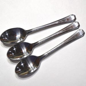 May include: Three stainless steel spoons arranged diagonally on a white surface. Each spoon has an oval bowl and a handle with a decorative, geometric end. The spoons reflect the surrounding light, creating highlights and shadows.