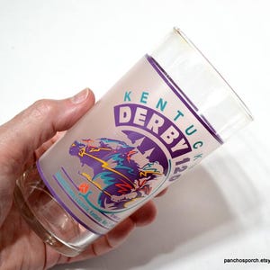 Puede incluir: Un vaso de vidrio transparente con un dise&ntilde;o esmerilado con las palabras "Kentucky Derby 120" en morado y verde. El dise&ntilde;o incluye un caballo y un jinete en movimiento. El vaso se sostiene en una mano.