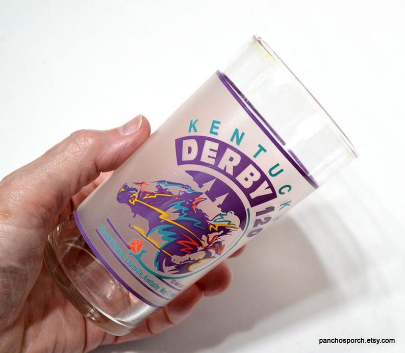 Puede incluir: Un vaso de vidrio transparente con un dise&ntilde;o esmerilado con las palabras "Kentucky Derby 120" en morado y verde. El dise&ntilde;o incluye un caballo y un jinete en movimiento. El vaso se sostiene en una mano.