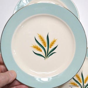 May include: A set of vintage ceramic plates. Each plate has a light blue border and a cream-colored center. The center of each plate features a design of wheat stalks in yellow and green. The plates have a gold trim.