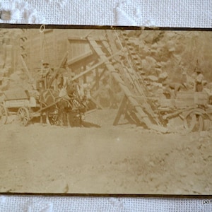 May include: A black and white vintage photograph of a horse-drawn wagon being pulled up a steep incline. The wagon is loaded with wood and has two men on board. The scene is likely from a logging operation in the early 1900s.