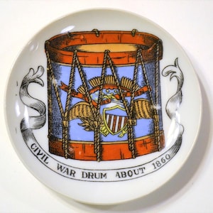 May include: A white ceramic plate featuring a Civil War drum design. The drum is depicted in orange, blue, and brown, with a shield and ribbon details. Text on the plate reads "Civil War Drum About 1860."