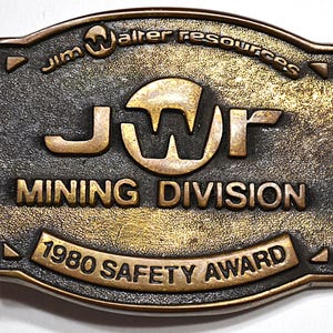 May include: A vintage bronze-colored metal belt buckle with the text "Jim Walter Resources" and "JWR MINING DIVISION 1980 SAFETY AWARD". The buckle has an oval shape with a textured surface and a dark patina.