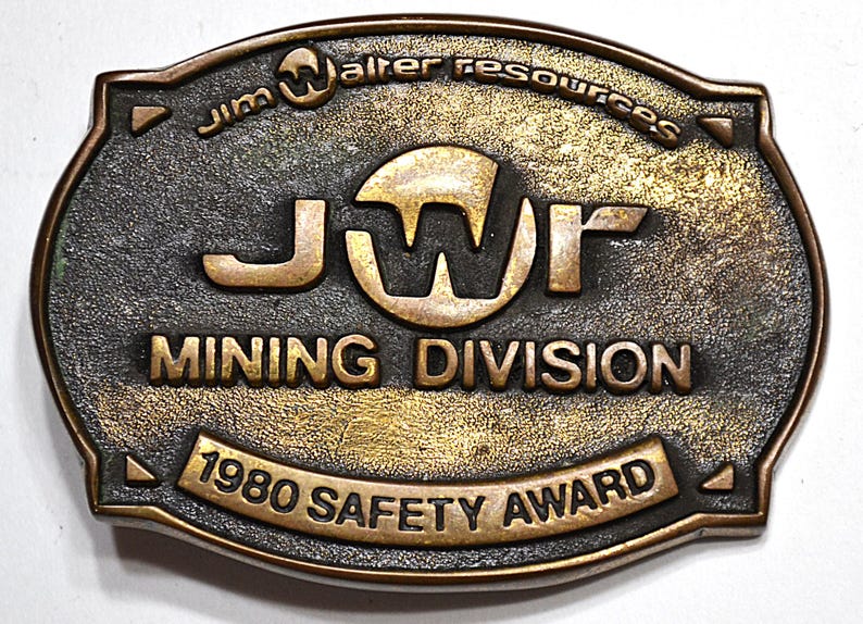 May include: A vintage bronze-colored metal belt buckle with the text "Jim Walter Resources" and "JWR MINING DIVISION 1980 SAFETY AWARD". The buckle has an oval shape with a textured surface and a dark patina.