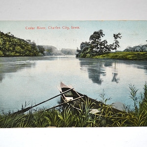 May include: A vintage postcard featuring a colourised illustration of the Cedar River in Charles City, Iowa. The scene depicts a small wooden rowboat with oars resting near a grassy riverbank, with trees lining the calm water under a pale sky.