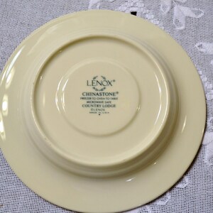 May include: Underside of a cream-colored Lenox Chinastone plate. The plate has a wide rim and a recessed center. Text on the plate reads "LENOX CHINASTONE FREEZER TO OVEN TO TABLE MICROWAVE SAFE COUNTRY LODGE GLENOS MADE IN U.S.A."