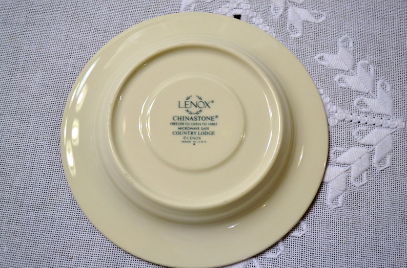 May include: Underside of a cream-colored Lenox Chinastone plate. The plate has a wide rim and a recessed center. Text on the plate reads "LENOX CHINASTONE FREEZER TO OVEN TO TABLE MICROWAVE SAFE COUNTRY LODGE GLENOS MADE IN U.S.A."