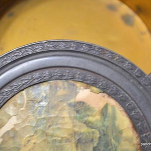 Op de afbeelding: Een antieke ronde metalen doos met een decoratieve rand en een landschapsafbeelding op het deksel. De binnenkant is goudgeel van kleur. Het deksel is gedeeltelijk open en onthult de binnenkant van de doos.