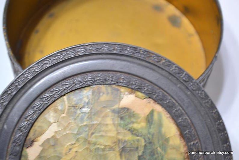 Op de afbeelding: Een antieke ronde metalen doos met een decoratieve rand en een landschapsafbeelding op het deksel. De binnenkant is goudgeel van kleur. Het deksel is gedeeltelijk open en onthult de binnenkant van de doos.