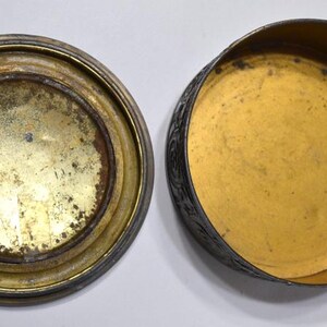 Op de afbeelding: Een open, antieke metalen doos met een deksel. Het deksel en de basis zijn cirkelvormig met een goudkleurig interieur en een aangetaste, verouderde uitstraling. De buitenkant van de doos is van donkerder, mogelijk zilverkleurig metaal.