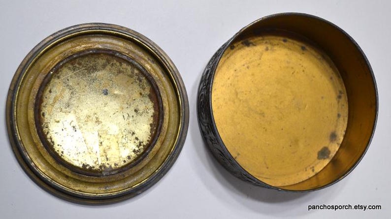Op de afbeelding: Een open, antieke metalen doos met een deksel. Het deksel en de basis zijn cirkelvormig met een goudkleurig interieur en een aangetaste, verouderde uitstraling. De buitenkant van de doos is van donkerder, mogelijk zilverkleurig metaal.
