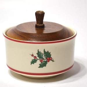 May include: A cream-colored ceramic sugar bowl featuring a red band around the top and bottom rim. The center displays a green holly leaf and red berry illustration. The bowl includes a circular dark brown wooden lid with a central knob handle.
