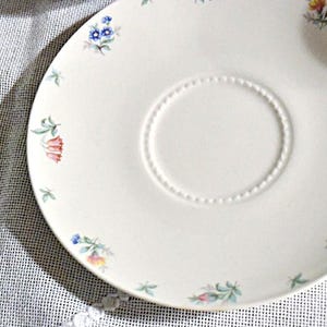 May include: A white ceramic saucer with a floral design. The saucer has a raised rim and a small depression in the center. The flowers are in shades of pink, blue, and green.
