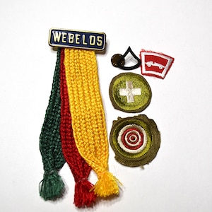 May include: A Webelos pin with three hanging braided ribbons in green, red, and yellow. Beside it are four embroidered patches: a black chevron, a red and white car emblem, a green circle with a white cross, and a green circle with a target.