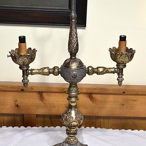 Puede incluir: Una l&aacute;mpara de mesa de dos luces estilo candelabro con acabado met&aacute;lico en tonos dorados y plateados. La l&aacute;mpara presenta un poste central vertical con un asa de anillo en la parte superior, dos brazos que se extienden hacia copas en forma de hoja.