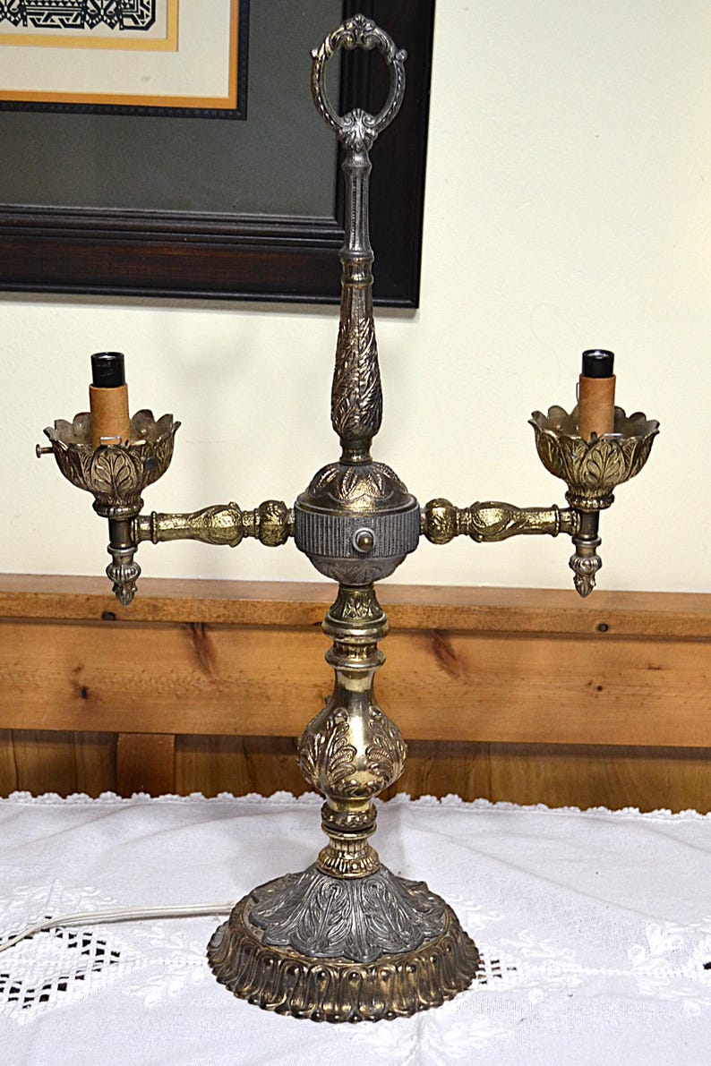 Puede incluir: Una l&aacute;mpara de mesa de dos luces estilo candelabro con acabado met&aacute;lico en tonos dorados y plateados. La l&aacute;mpara presenta un poste central vertical con un asa de anillo en la parte superior, dos brazos que se extienden hacia copas en forma de hoja.