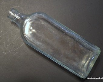 May include: A clear, rectangular glass bottle with a rounded neck and a flat base. The bottle is empty and has a slightly bluish tint. The surface shows some wear and tear. The bottle is lying on a dark surface.