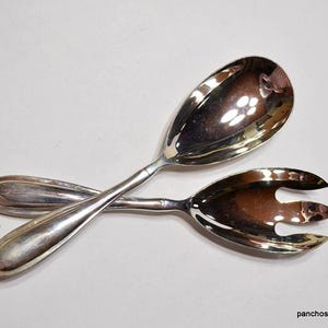 May include: A pair of silver serving utensils, a spoon and a fork, are crossed against a white background. The spoon has a large, oval bowl, and the fork has three tines. The utensils are reflective.