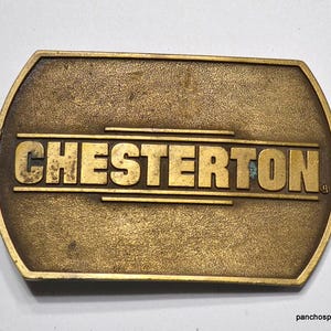 May include: A vintage brass belt buckle with the name "CHESTERTON" in bold, raised letters. The buckle has a rectangular shape with rounded edges and a weathered, antique finish. The text is centered on the buckle.