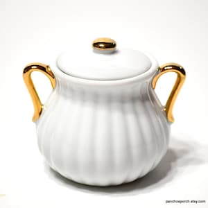 May include: White ceramic sugar bowl with a lid and gold-colored accents. The bowl has a ribbed design and two gold handles. The lid also has a gold knob. The sugar bowl is on a white background.