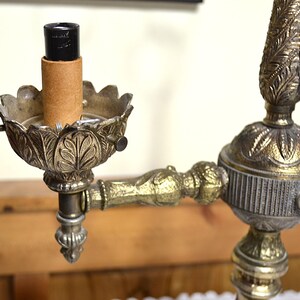 Puede incluir: Una l&aacute;mpara de metal en tono lat&oacute;n con una arandela decorativa de hojas, un casquillo central negro y una funda de cart&oacute;n. La estructura incluye un brazo decorativo y una columna central con detalles met&aacute;licos intrincados y texturizados.