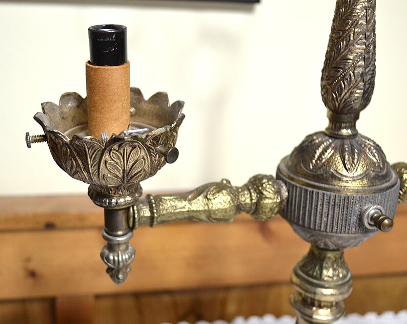 Puede incluir: Una l&aacute;mpara de metal en tono lat&oacute;n con una arandela decorativa de hojas, un casquillo central negro y una funda de cart&oacute;n. La estructura incluye un brazo decorativo y una columna central con detalles met&aacute;licos intrincados y texturizados.