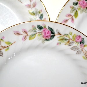 May include: White porcelain plates with a gold rim and a floral design featuring pink roses, green leaves, and pink accents. The plates have a slightly textured surface and are arranged in a close-up shot.