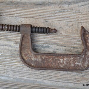 May include: A rusty metal C-clamp with a threaded handle. The clamp is open and ready to be used.