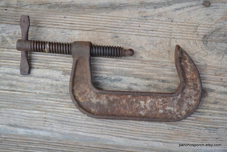 May include: A rusty metal C-clamp with a threaded handle. The clamp is open and ready to be used.