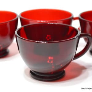 May include: Four ruby red glass teacups with handles. The cups have a rounded bowl shape and a small, circular base. The interior of the cups is a bright red, and the exterior is a darker, translucent red. The cups are arranged on a white surface.