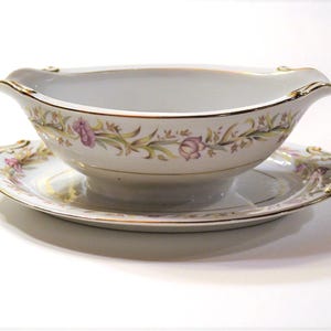 May include: A white porcelain gravy boat and matching saucer. The set features a floral design with pink and green accents, and gold trim. The gravy boat has a curved handle on each side.