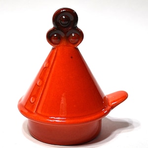 May include: A vibrant orange ceramic tagine lid with a conical shape and a small handle. The lid features a decorative, dark brown, three-circle design on top. The tagine lid is designed to fit on a matching base.