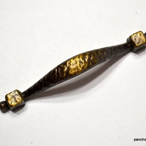 May include: A bronze-toned cabinet pull with a textured, hammered finish. The center section is wider and lighter in color, with square end pieces and rounded accents. A decorative hardware piece.