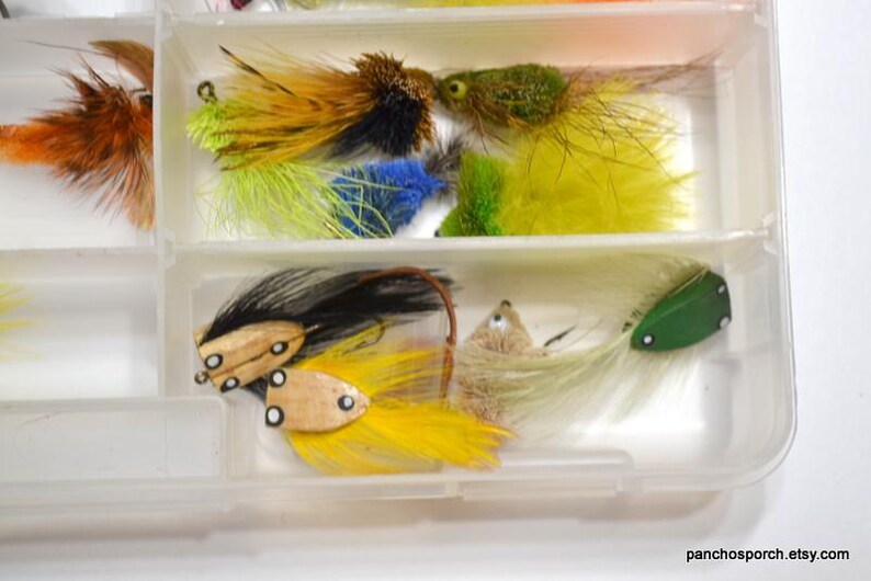 May include: A clear plastic tackle box filled with colorful fishing flies. The flies feature various colors, including yellow, green, orange, and black, with feather and fur details. The flies are arranged in separate compartments.