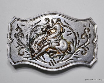 May include: Silver-toned metal belt buckle with a cowboy riding a bucking bronco. The buckle has an oval shape with decorative scrollwork around the edges and a gold-colored accent on the cowboy and horse. The buckle is slightly distressed.