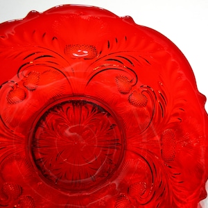 May include: A vibrant red glass bowl featuring a scalloped edge and an intricate embossed pattern of thistle flowers and curved stems. The center of the bowl displays a starburst design, while the translucent glass catches light throughout.
