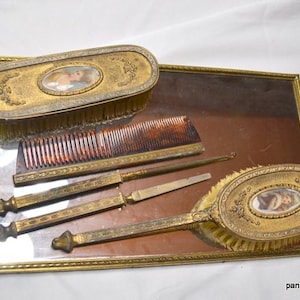 May include: A vintage gold-toned vanity set with a mirror, two brushes, a comb, and two metal tools. The set is decorated with ornate details and floral motifs.