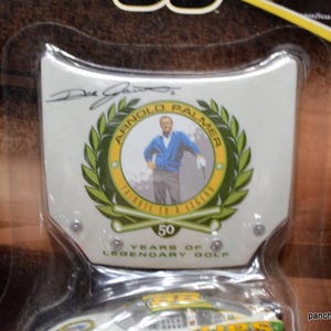 May include: A collectible item featuring a portrait of Arnold Palmer within a green laurel wreath. The design includes the text "Arnold Palmer" and "Tribute to a Legend". The item commemorates 50 years of legendary golf.