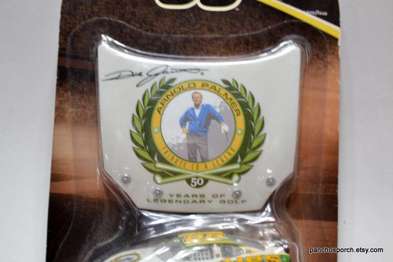 May include: A collectible item featuring a portrait of Arnold Palmer within a green laurel wreath. The design includes the text "Arnold Palmer" and "Tribute to a Legend". The item commemorates 50 years of legendary golf.