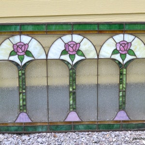 Op de afbeelding: Een glas-in-loodraam met drie gebogen panelen met roze rozen en groene bladeren. Het raam is omlijst met bruin en groen.