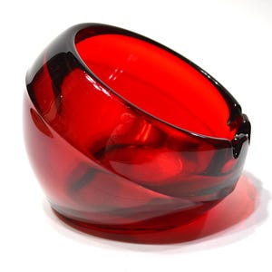 May include: A vibrant red glass ashtray with a smooth, rounded, and asymmetrical bowl shape. The thick, translucent glass features a deep crimson hue with darker red swirls integrated into the structure, including a small notch on the rim.