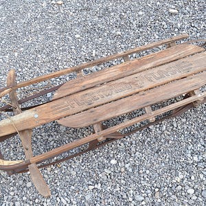 May include: Vintage wooden sled with the text "Fleetwing Flash" on the seat. The sled has wooden slats, metal runners, and a curved front. The sled is weathered, showing signs of age and use. A classic winter recreational item.