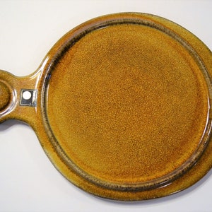 May include: A round, ceramic serving plate with a handle. The plate is a mottled, golden-brown color with a textured surface. A dark brown band encircles the edge. The handle has a circular opening. A small, white label is affixed to the handle.
